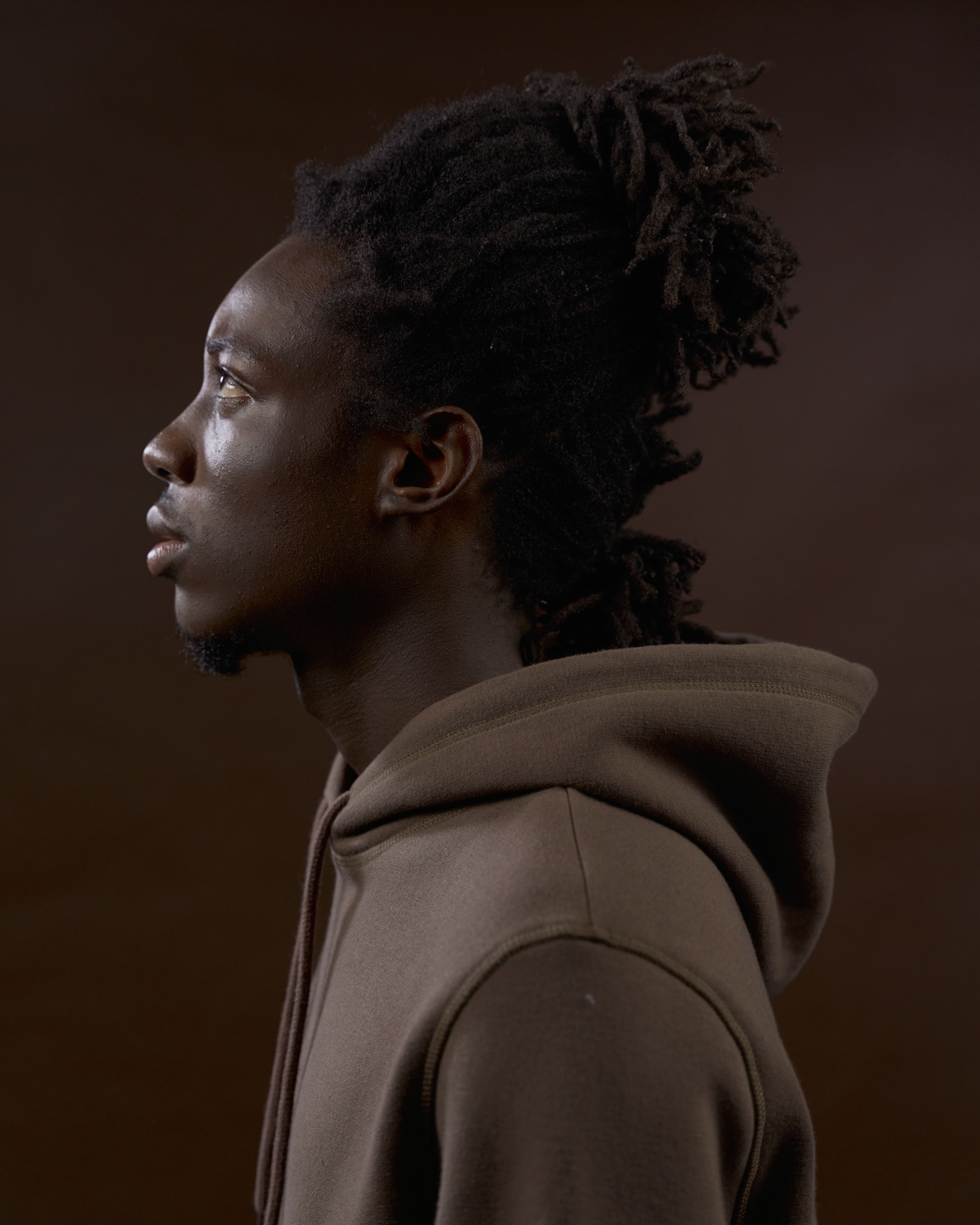 Side view portrait of a black man with afro hair in a brown hoodie against a dark background.