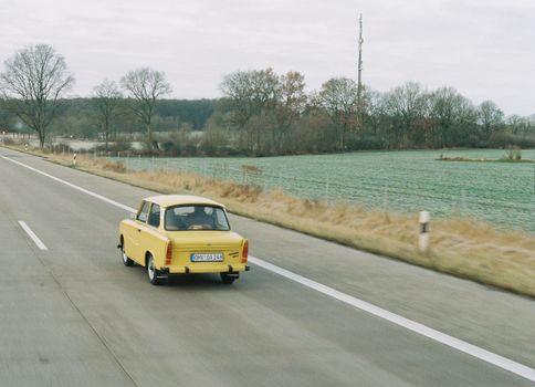 A classic yellow Trabant car travels on an empty road through a scenic countryside landscape.