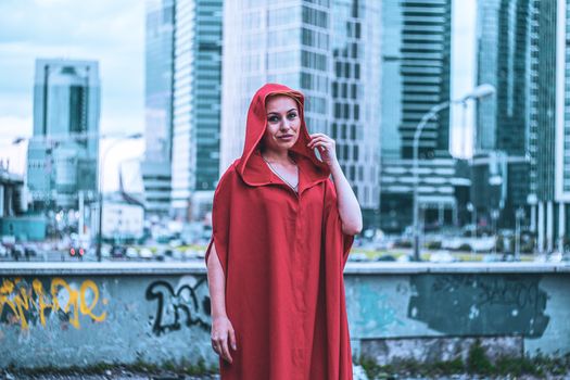 A woman stands in a red cloak against a modern cityscape background, evoking fashion and mystery.