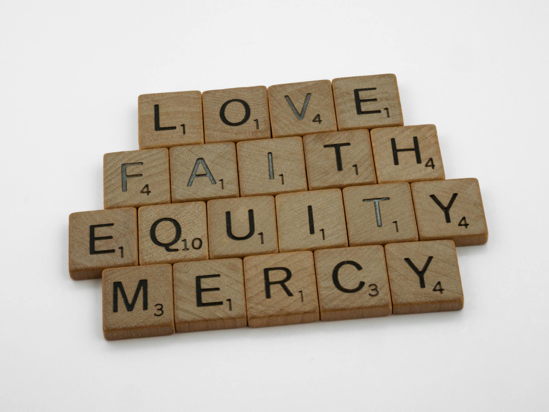 Wooden Scrabble tiles arranged to spell 'Love', 'Faith', 'Mercy', 'Equity' on a white background.