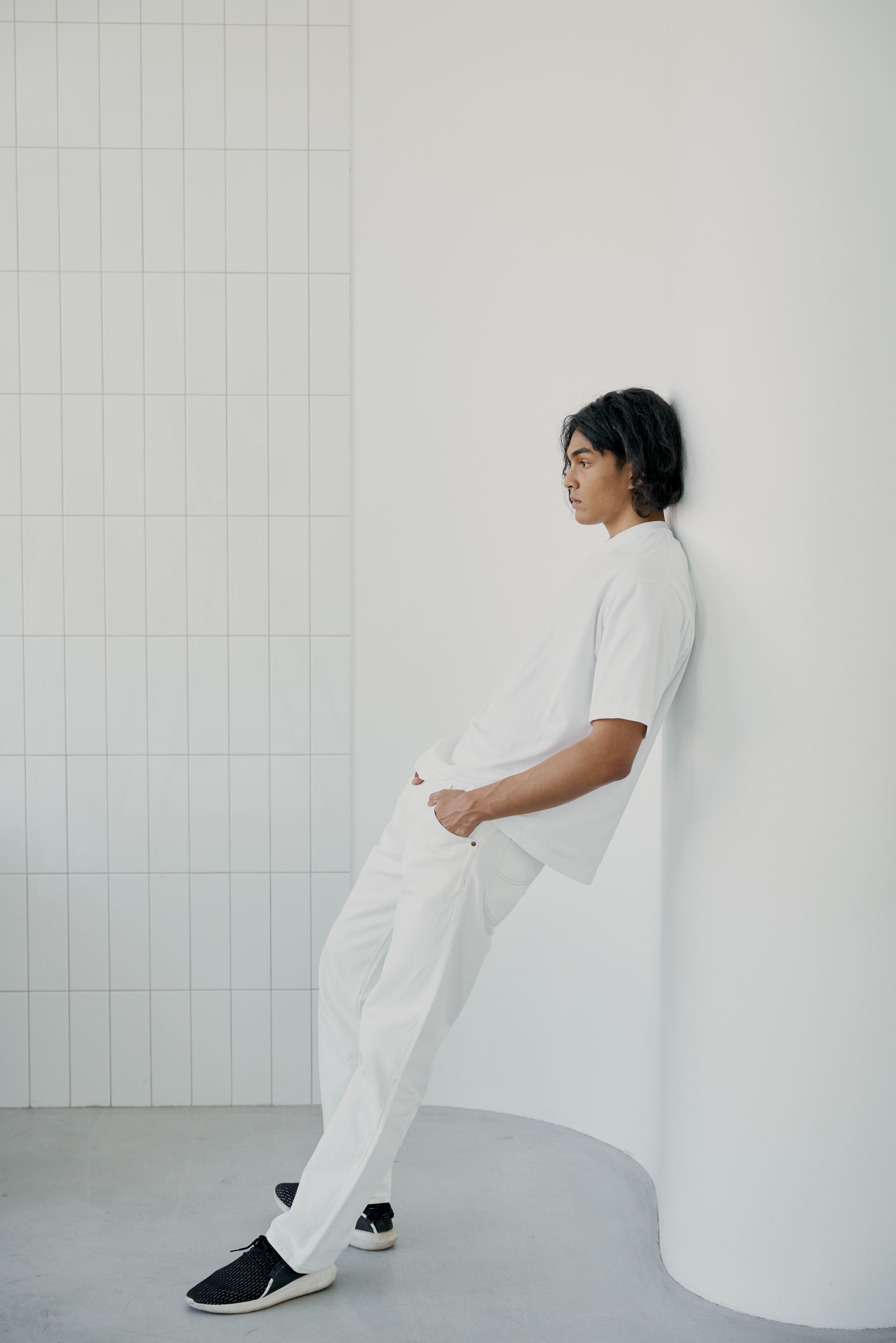 Stylish man in white outfit leaning against a minimalist white curved wall, indoor setting.