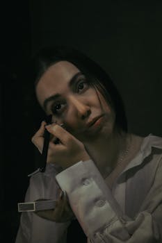 A woman applies makeup under soft lighting, creating a moody atmosphere.