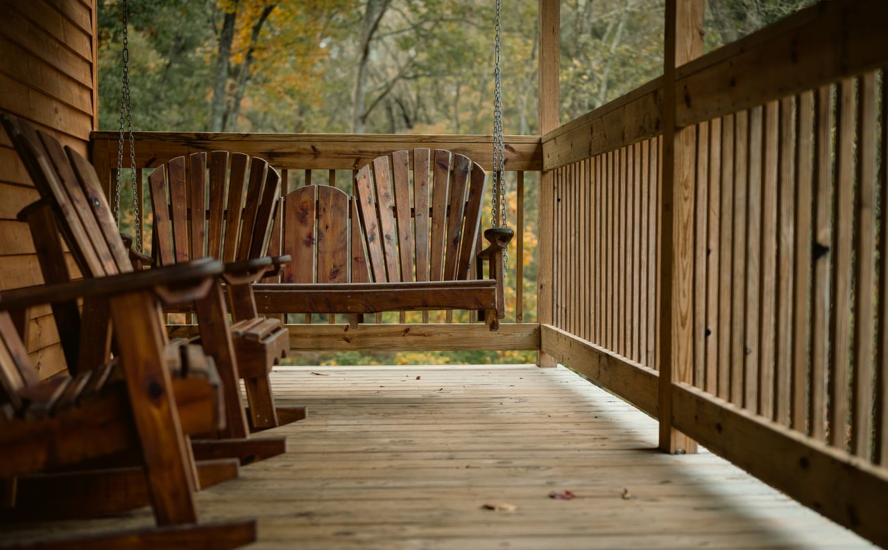 Wooden porch swing and Adirondack rocking chairs on a cabin porch with autumn trees behind the railing