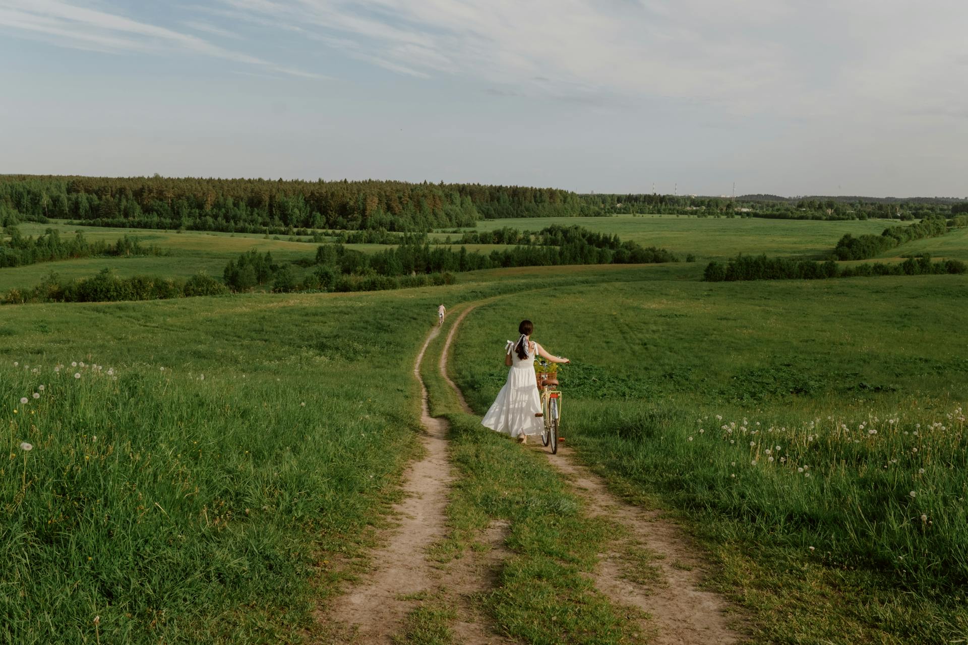 Woman in white dress walking a bicycle on a dirt path through a scenic countryside landscape.