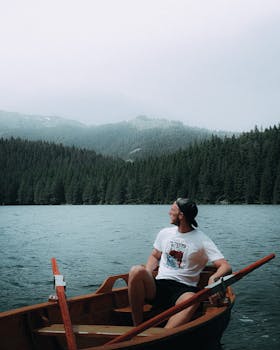 A man rowing a boat on a tranquil mountain lake surrounded by forested hills and fog.