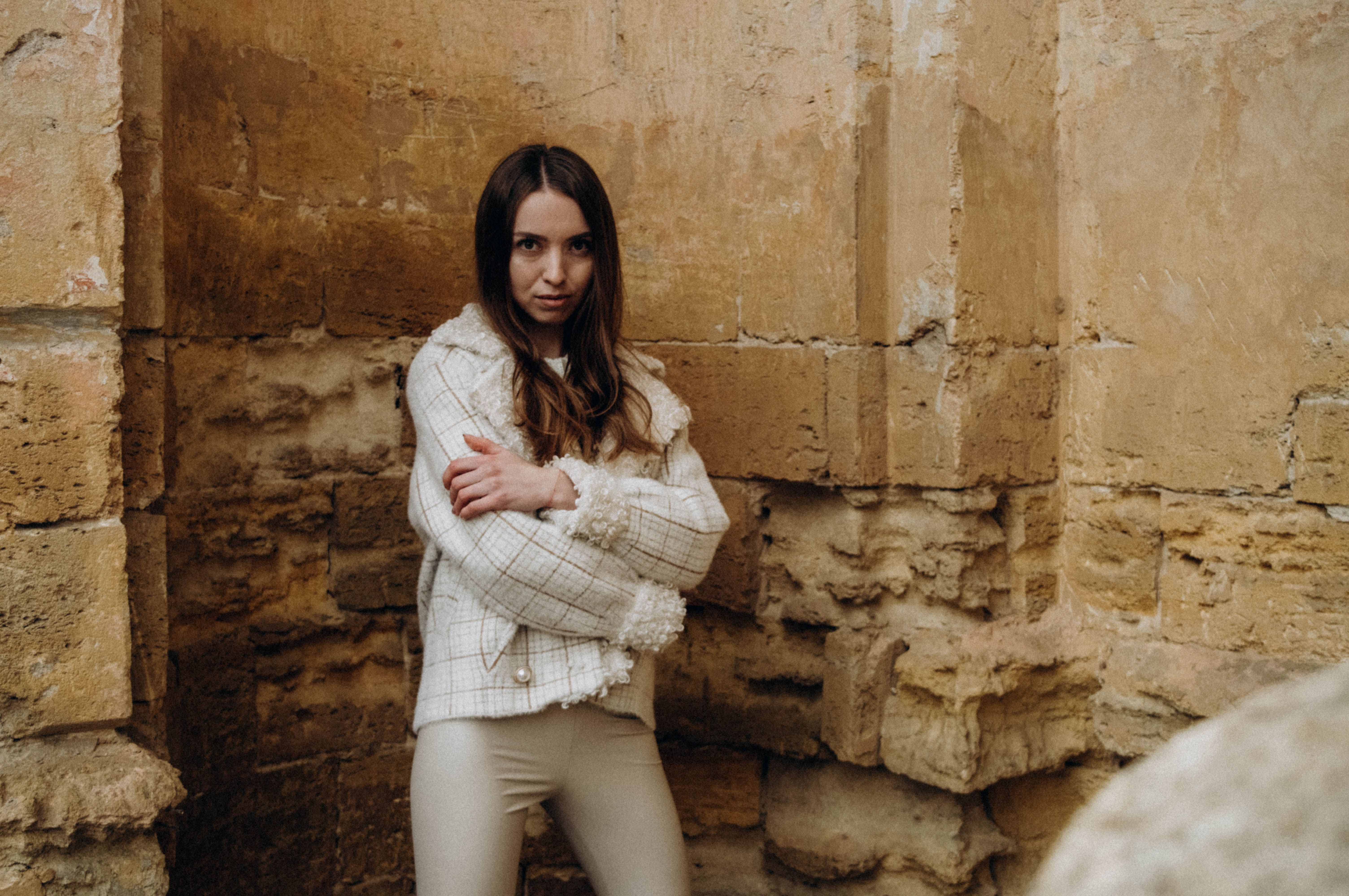 A woman wearing a cream tweed jacket with fringe trim and tonal cream trousers against a stone wall, showing tonal color strategy for tweed outfits