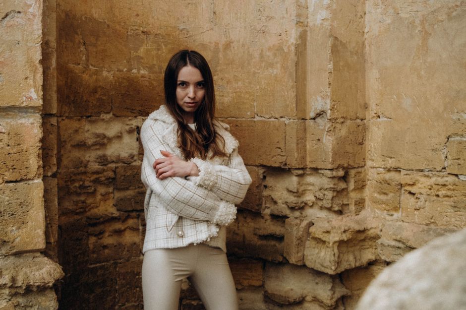 A woman wearing a cream tweed jacket with fringe trim and tonal cream trousers against a stone wall, showing tonal color strategy for tweed outfits