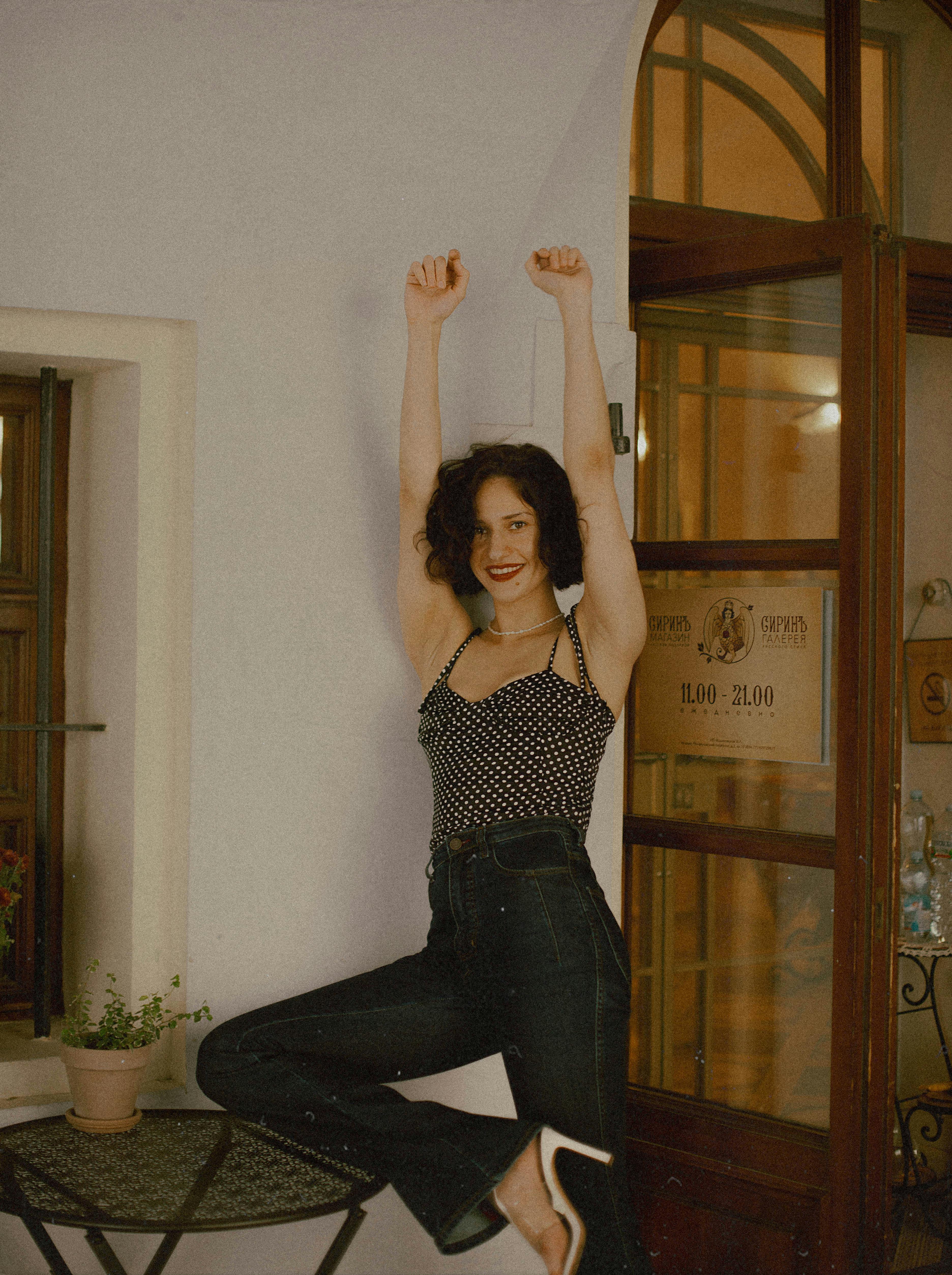 A joyful woman poses with arms raised, smiling in a stylish indoor setting.
