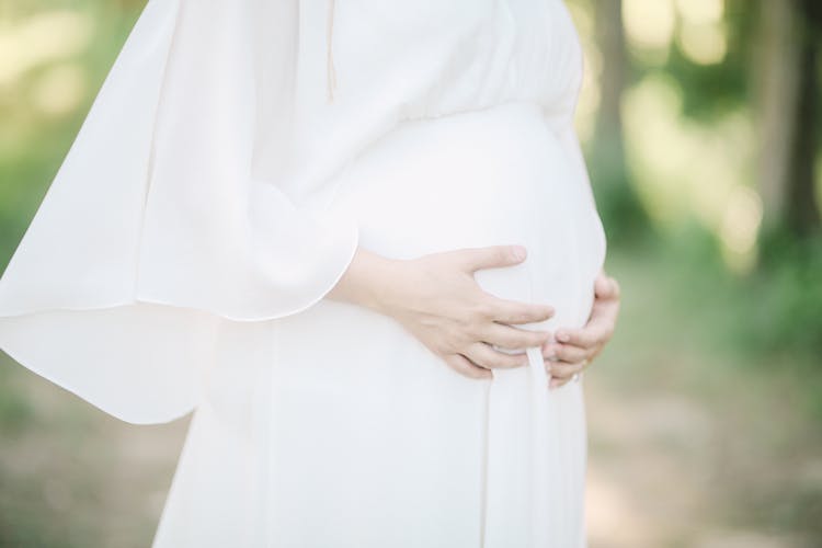 Unrecognizable Pregnant Woman In White Dress Holding Her Hands On Stomach