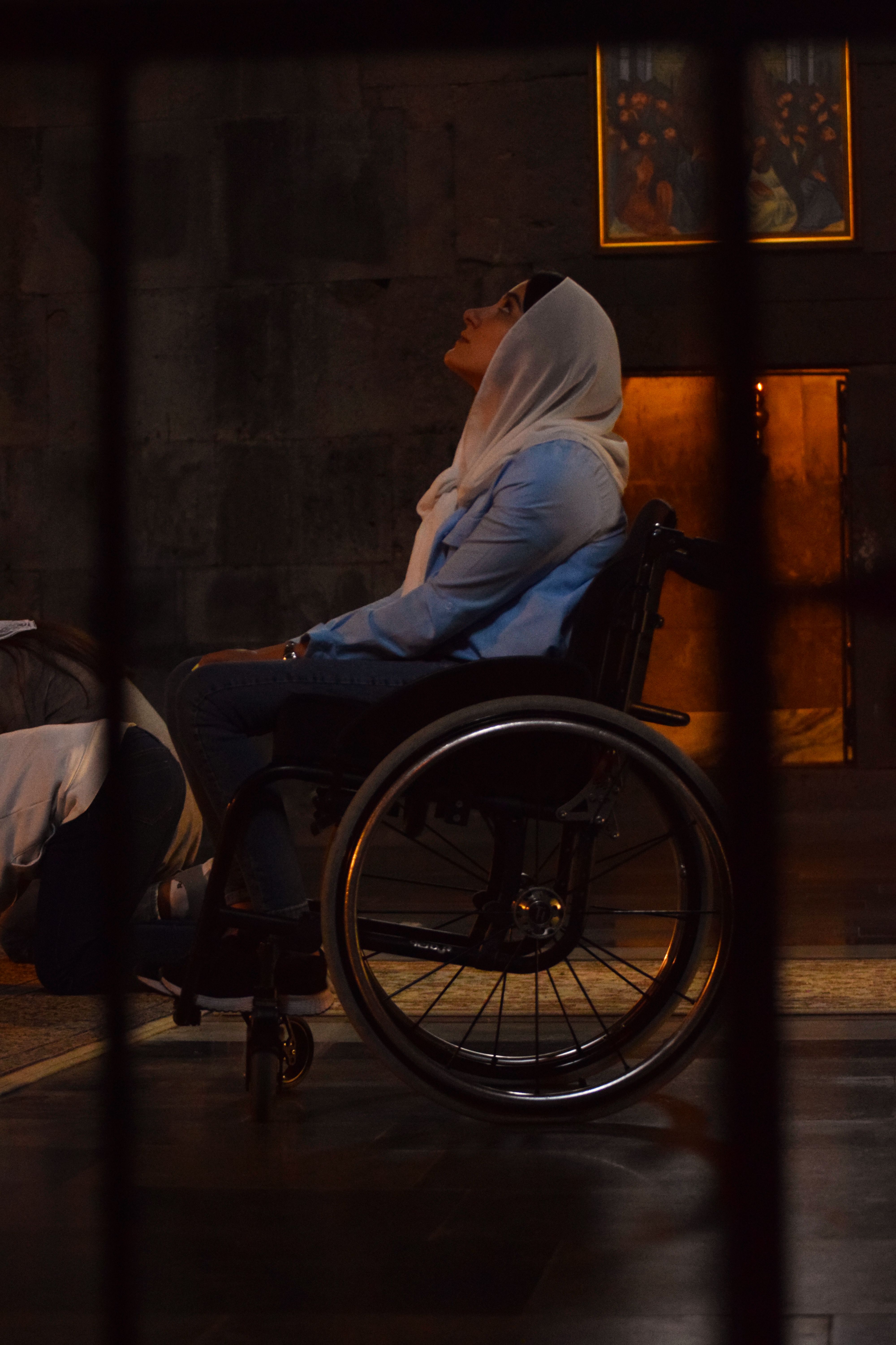 Woman in a wheelchair in prayer, inside a dimly lit religious setting.