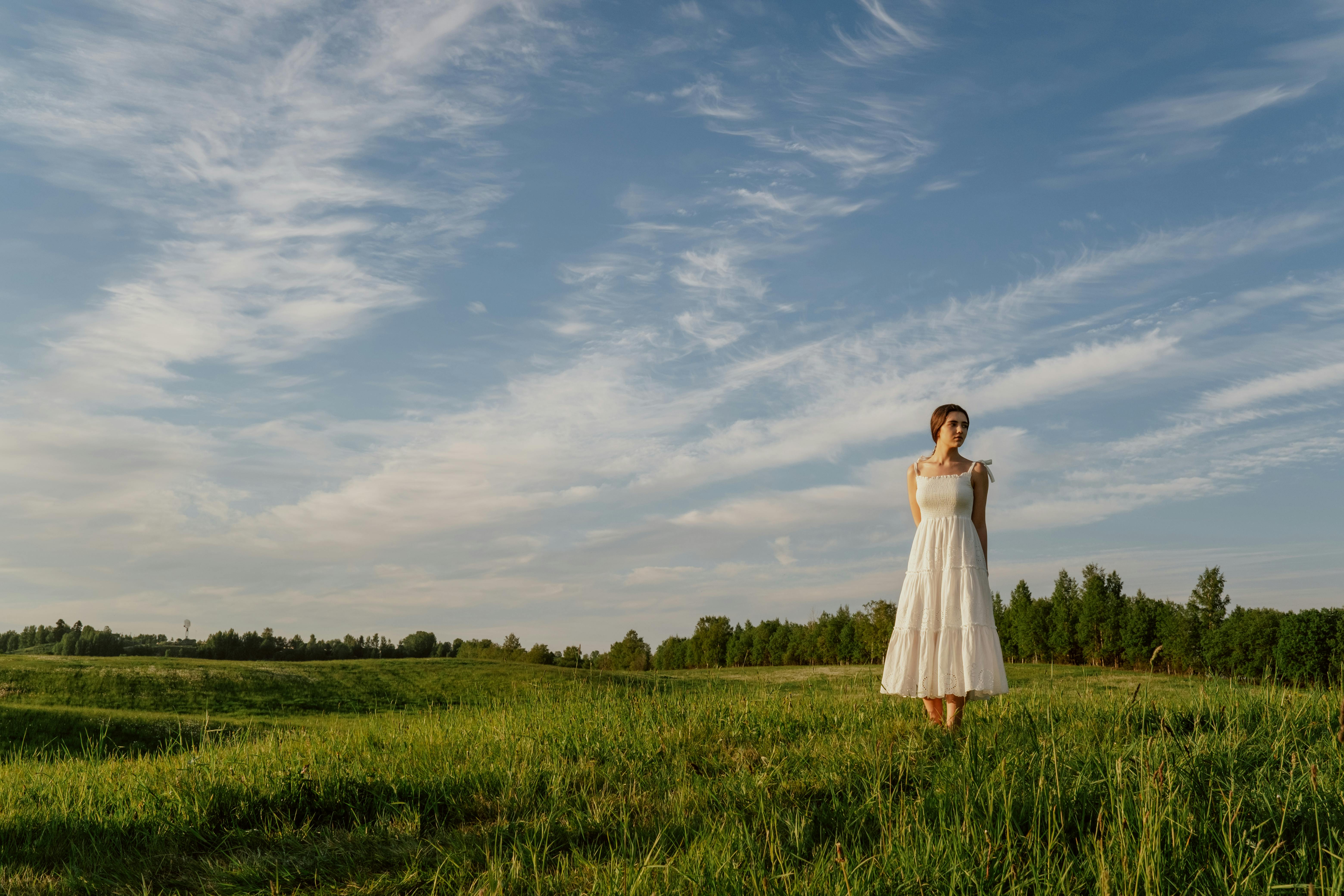 A woman in a white boho dress stands in a sunlit meadow under a clear sky.