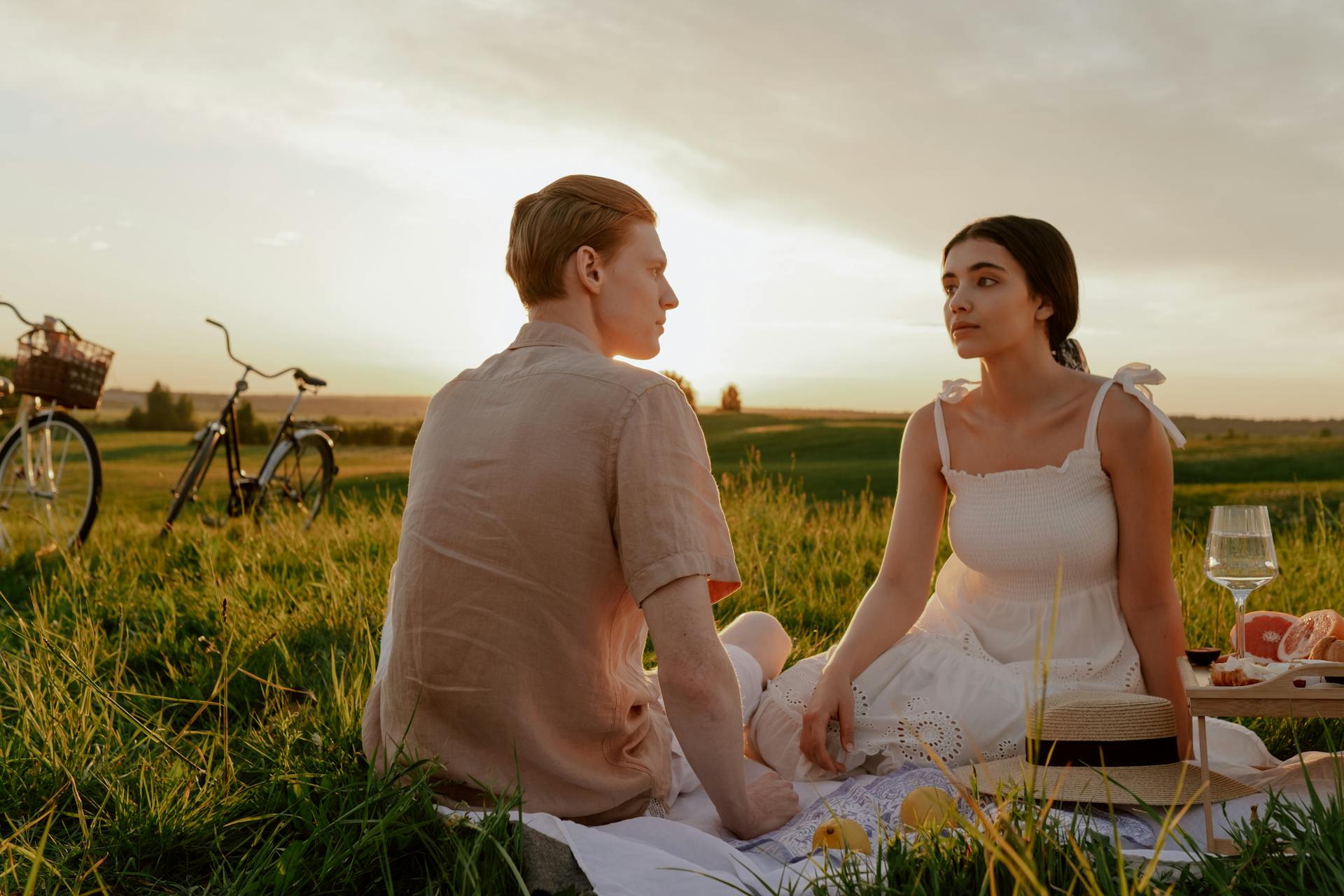 Couple enjoying a romantic picnic in a summer meadow at sunset with bicycles nearby.