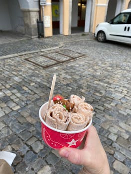 Hand holding rolled ice cream dessert in a red paper bowl on a cobblestone street, outdoors.