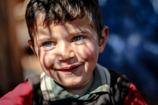 A young boy with blue eyes smiles warmly. Outdoor setting with sun casting shadows.