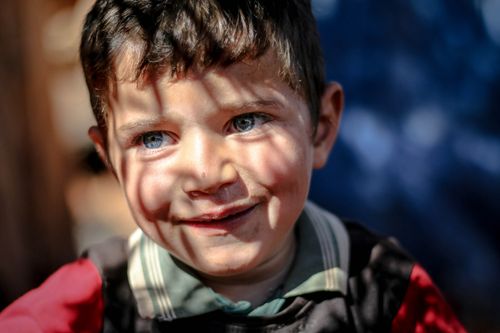 Free A young boy with blue eyes smiles warmly. Outdoor setting with sun casting shadows. Stock Photo