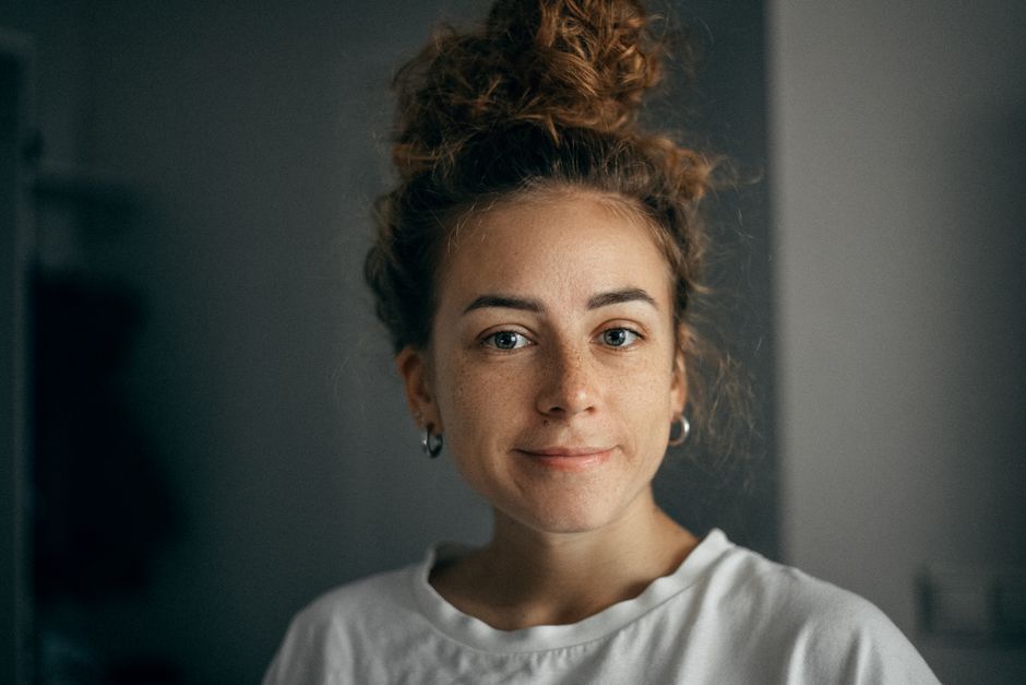 Portrait of a smiling woman with curly hair and freckles in soft light, wearing a white t-shirt indoors.