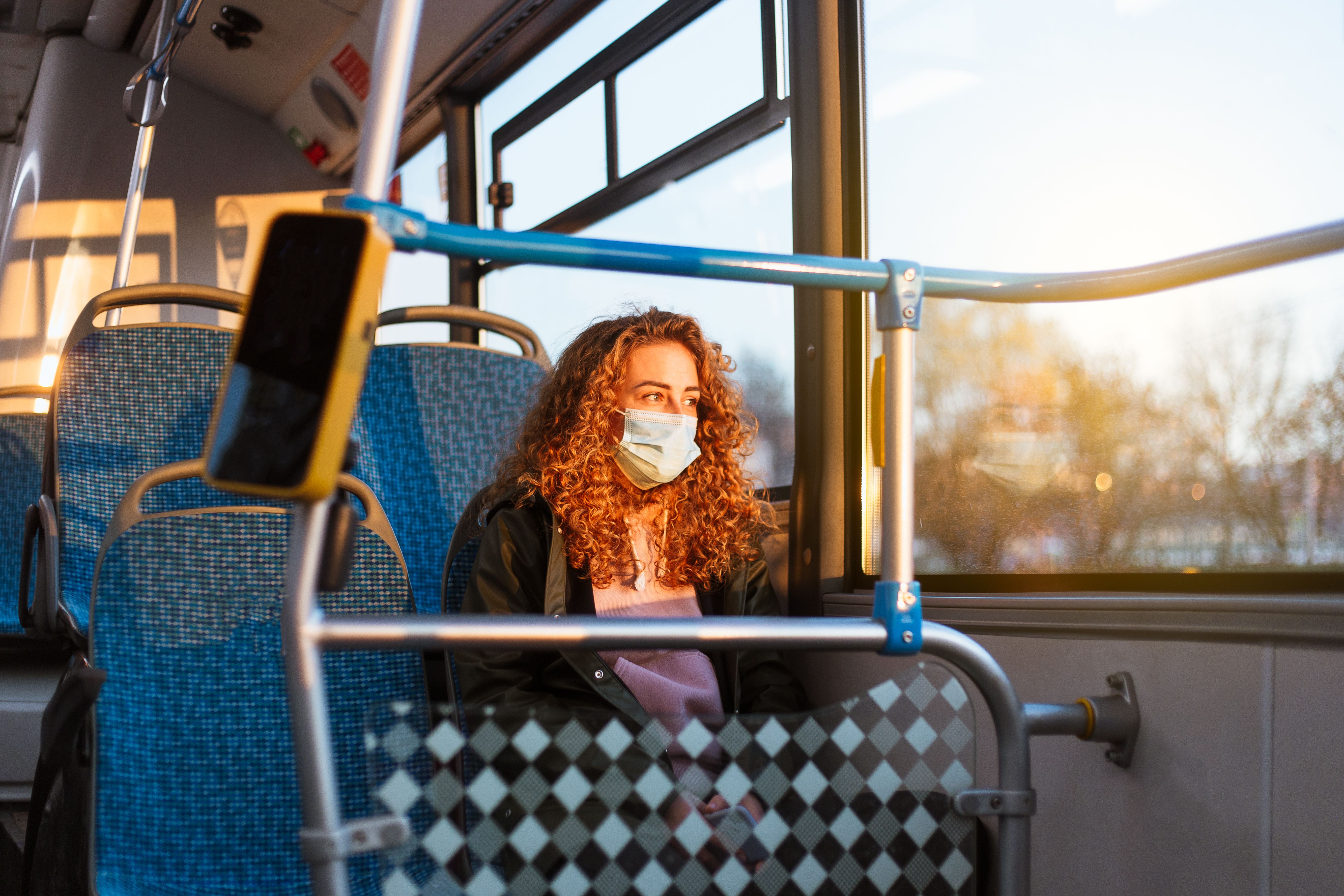 A woman with curly red hair wearing a face mask sits on a bus, gazing out the window during sunset.