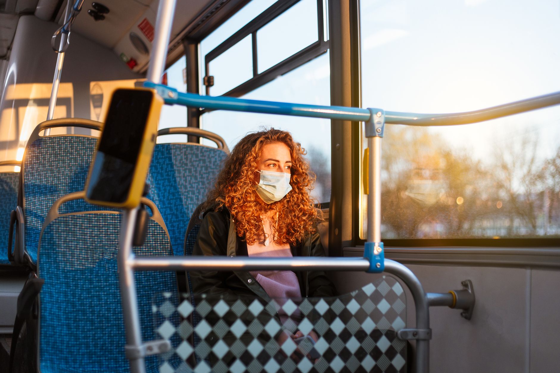 A woman with curly red hair wearing a face mask sits on a bus, gazing out the window during sunset.