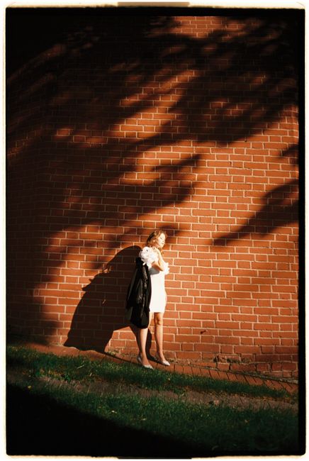 A woman in a white dress stands stylishly against a brick wall with dramatic shadows.