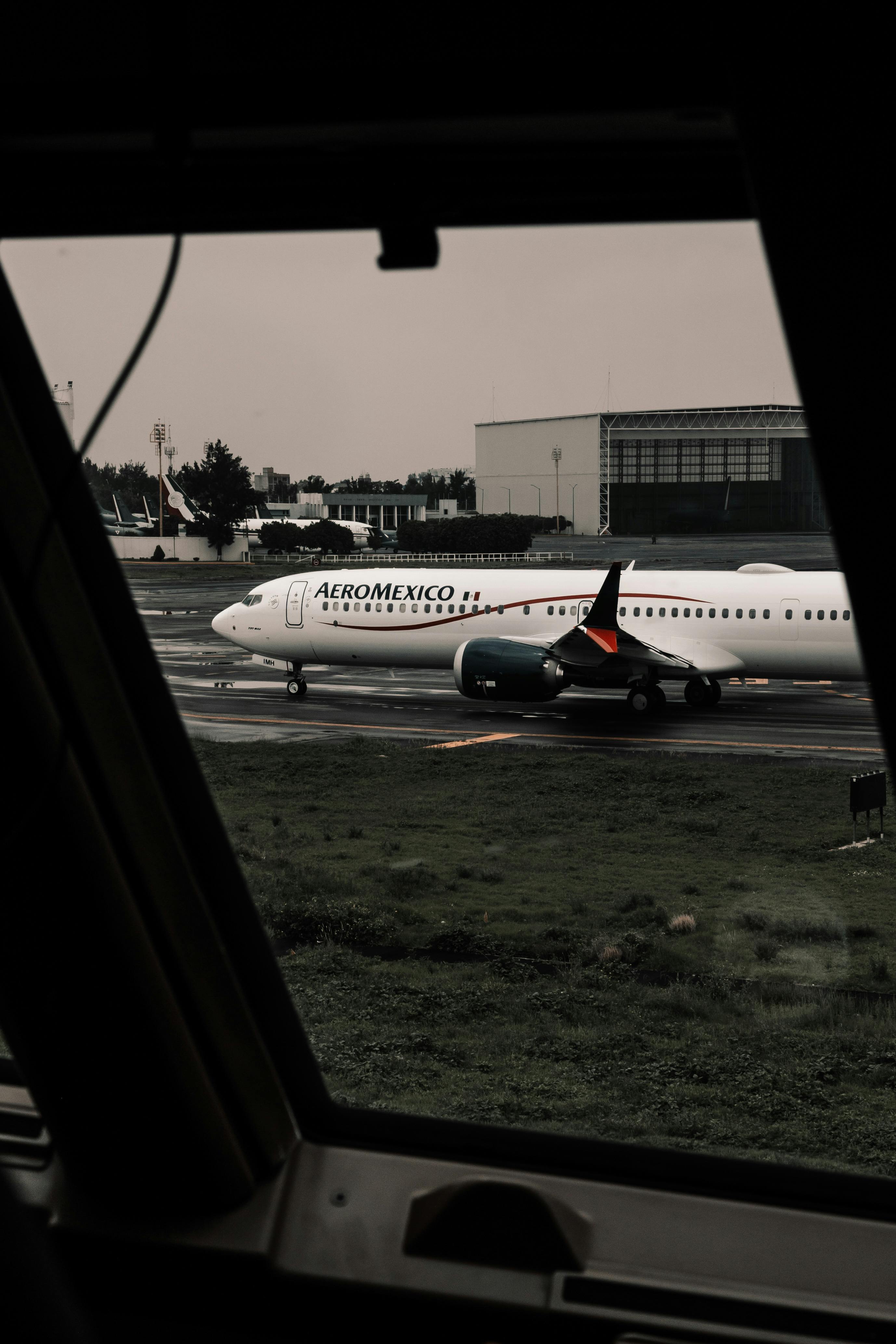 Free Aeromexico airplane photographed through a window, showcasing airport runway and surrounding structures. Stock Photo