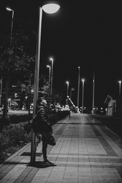 Grayscale Photo of Man Leaning on Street Lamp