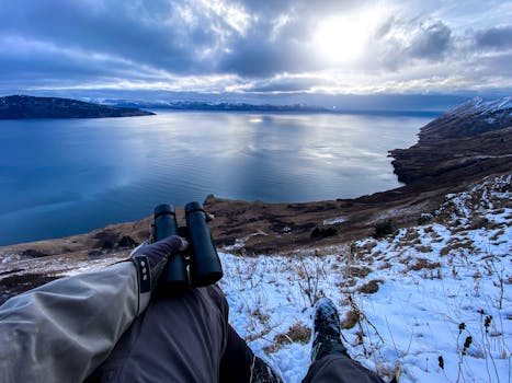 A person holding binoculars overlooking a snowy coastline with mountains and sea at dusk.