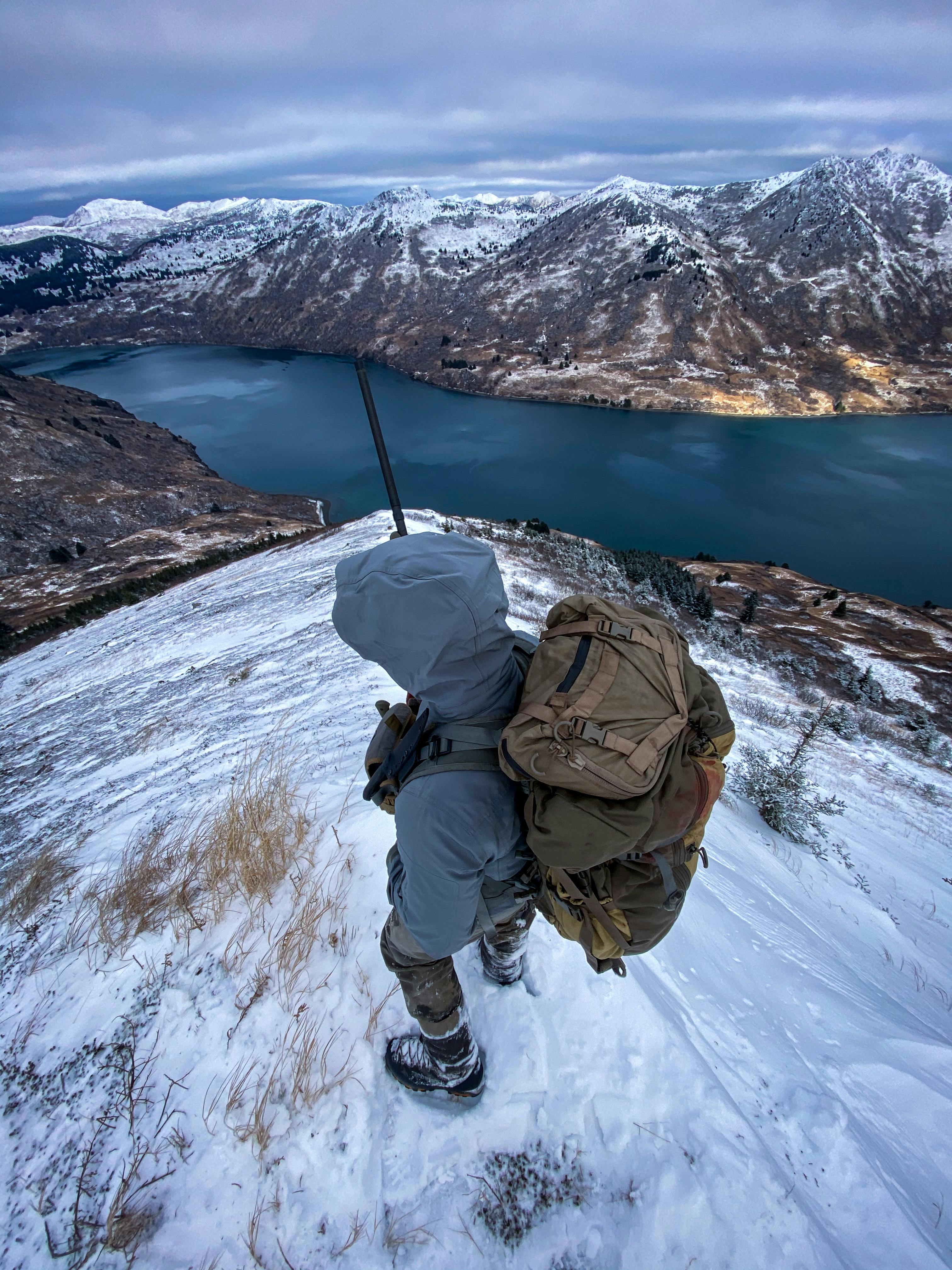Hiker with backpack on a snowy mountain overlooking a serene lake and mountain range.