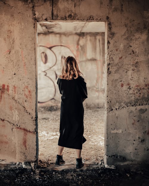 Blond Woman Wrapped in Black Coat Standing in Door of Ruined Building