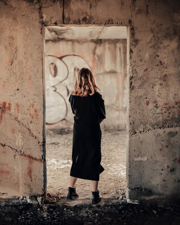 Blond Woman Wrapped In Black Coat Standing In Door Of Ruined Building