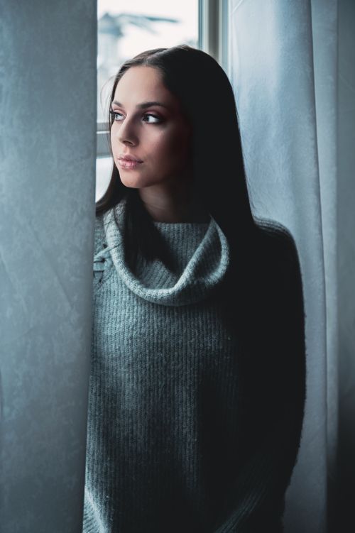 Free Portrait of a young woman in a cozy gray sweater, gazing thoughtfully out a window. Stock Photo