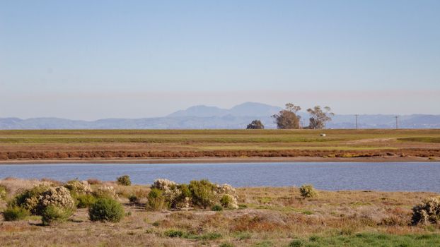 A tranquil rural scene featuring a calm river, open fields, and distant mountains under a clear sky.