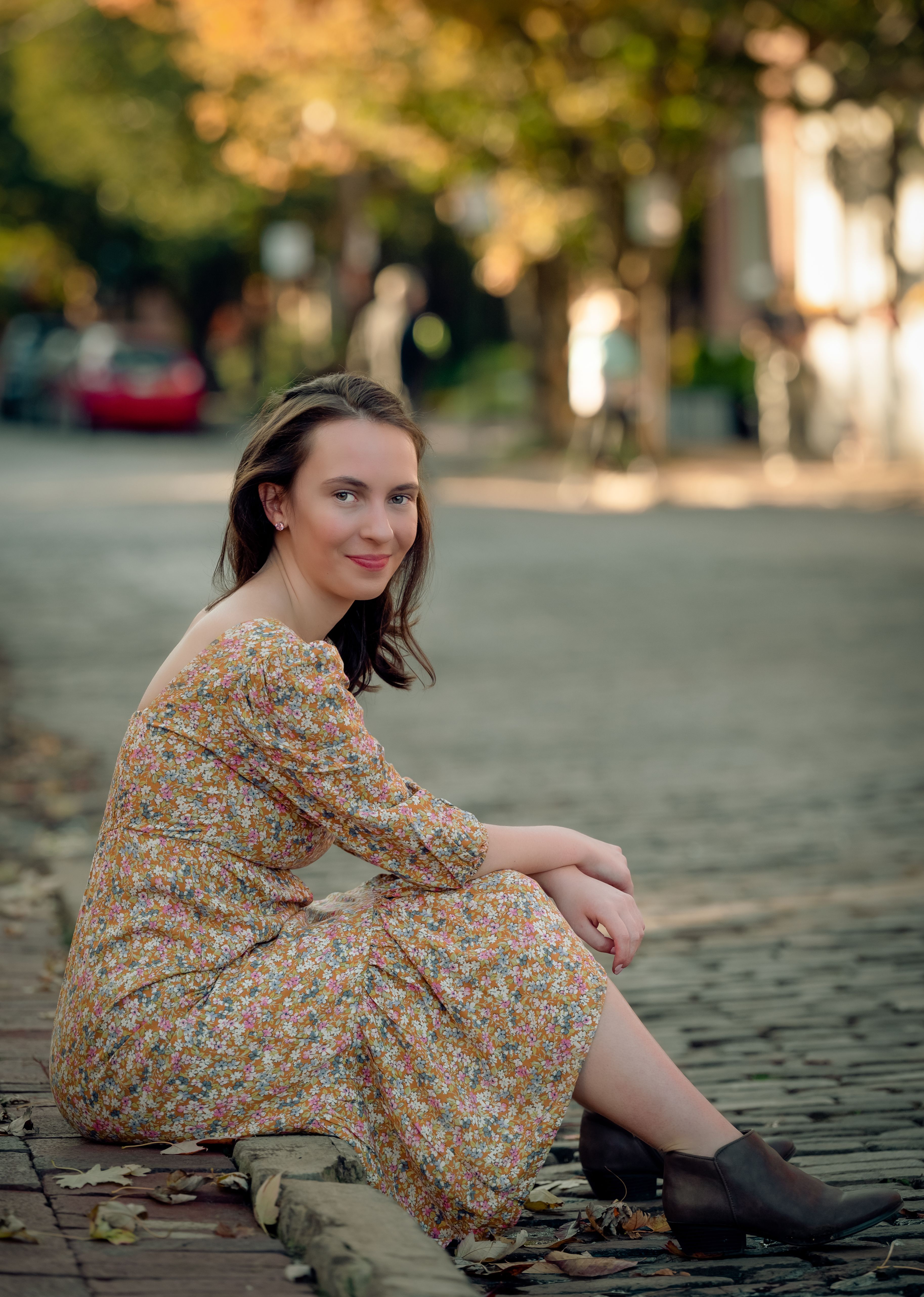 Elegant young woman wearing a floral dress sits gracefully on a cobblestone street in an urban setting.