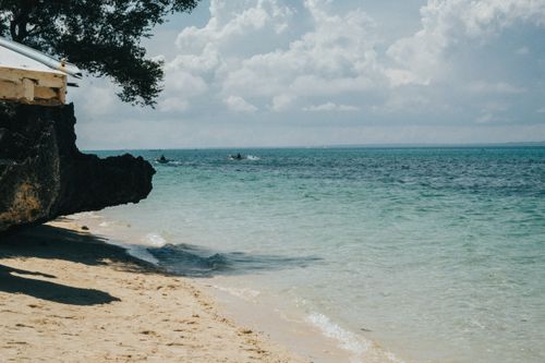 Free Idyllic beach scene with clear turquoise water and lush foliage under a partly cloudy sky. Stock Photo