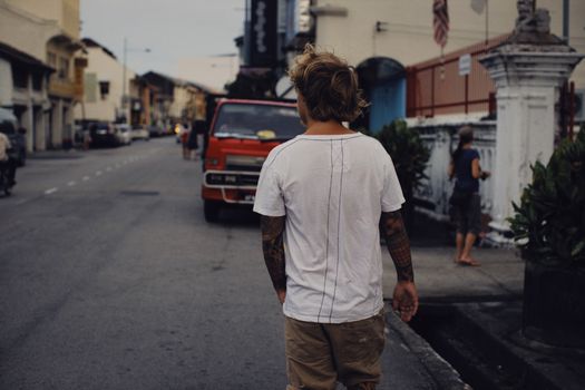 A tattooed man in a white shirt walks down a city street, showcasing urban lifestyle.