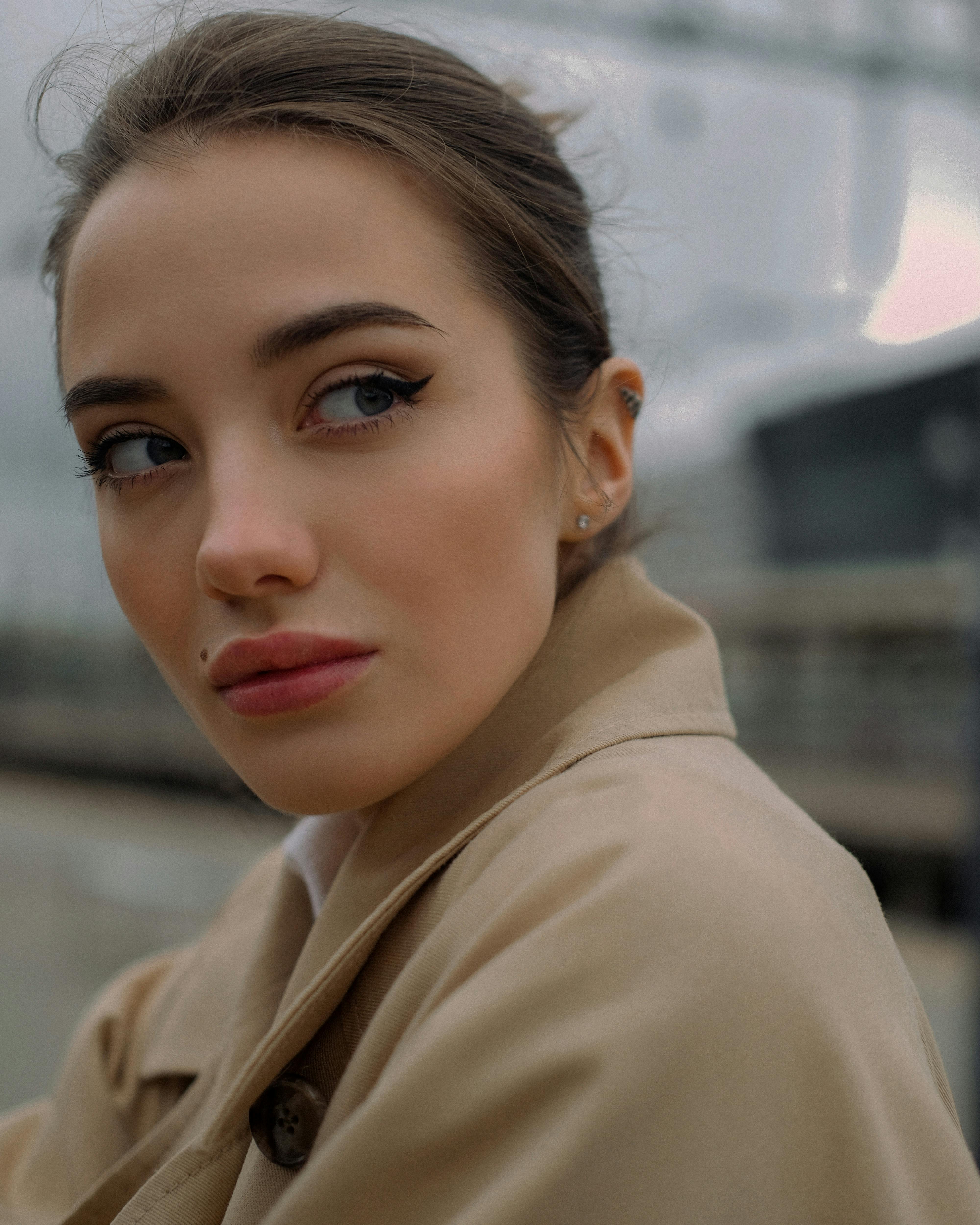 A woman in a beige trench coat gazes thoughtfully outdoors, exuding elegance and style.