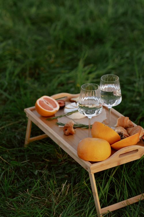 Free A wooden tray with fruits, croissants, and drinks set on lush grass for a refreshing picnic. Stock Photo