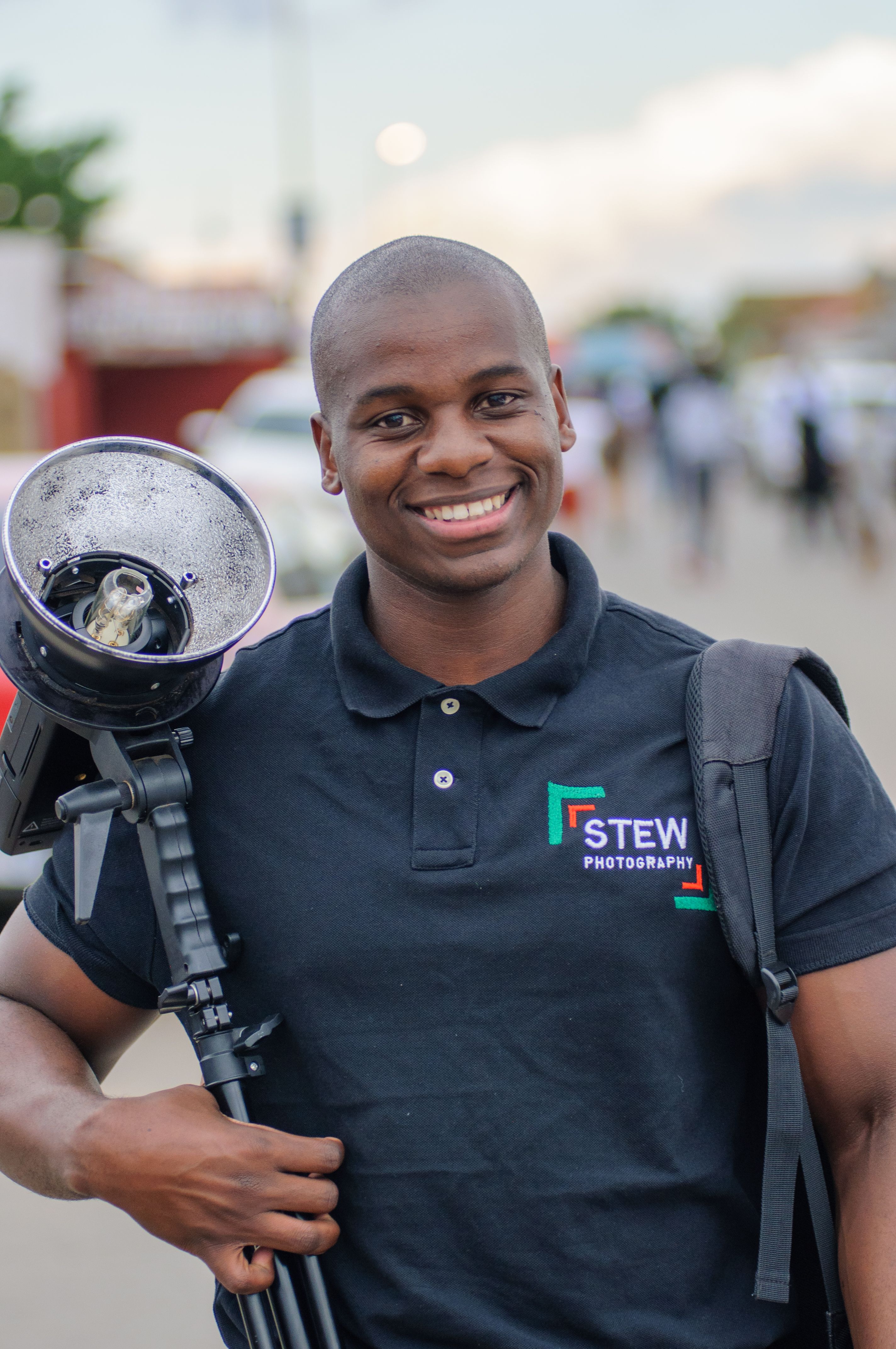 African American photographer smiling while holding equipment outdoors, wearing a polo shirt.