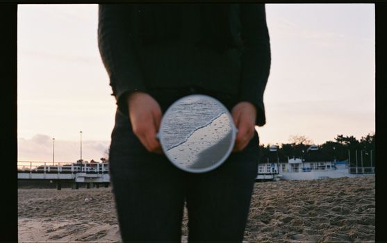 A person holding a round mirror on a sandy beach reflecting the shoreline at dusk.