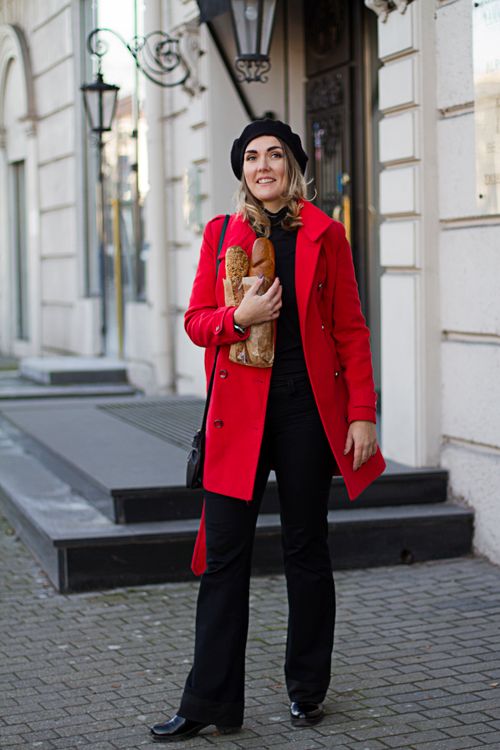 Free Fashionable woman in red coat holding baguettes, smiling outdoors in an urban setting. Stock Photo