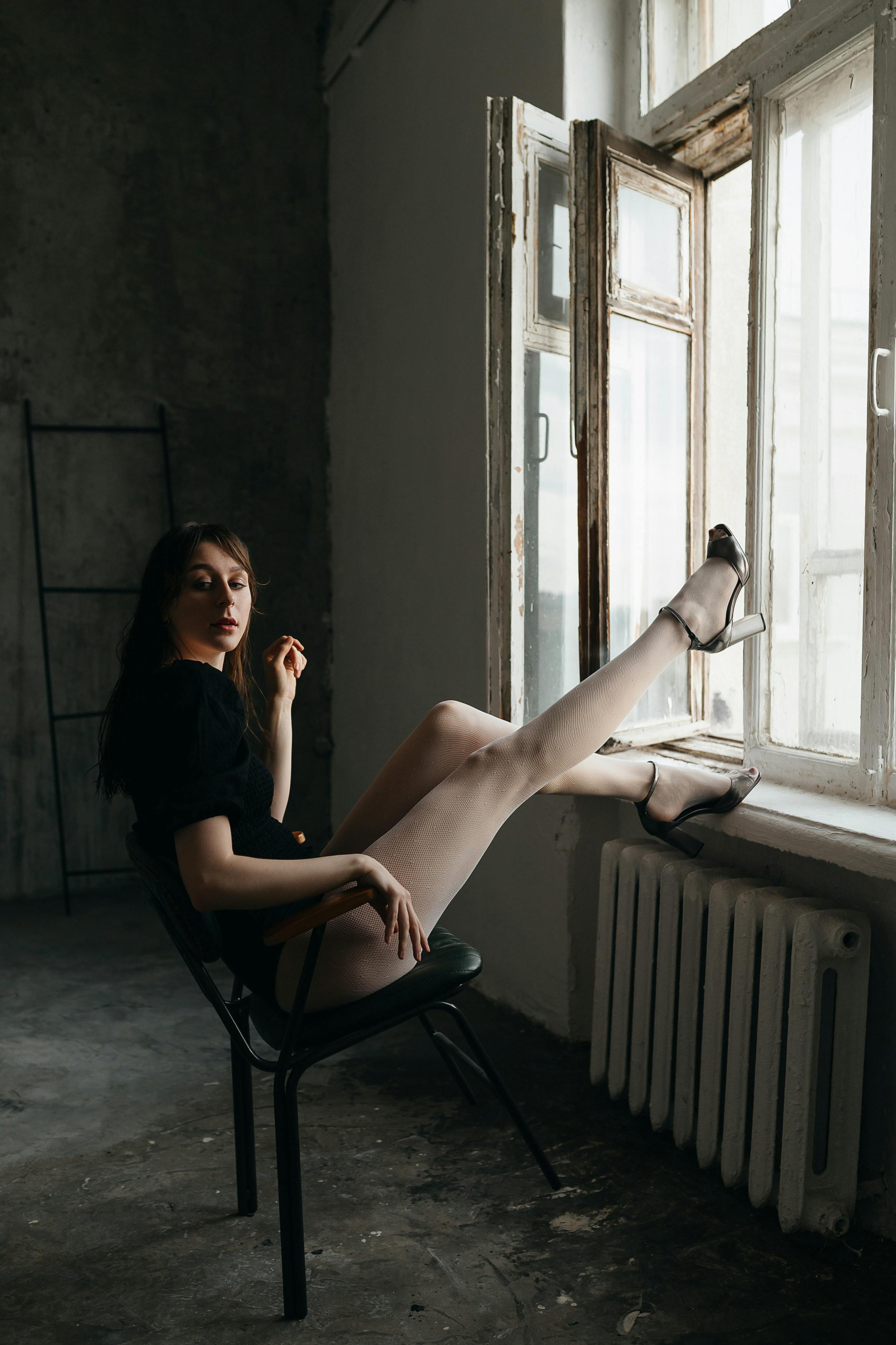 Woman in high heels sits on a chair, legs outstretched to a windowsill, creating a chic and relaxed ambiance.