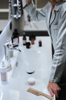 A woman in pajamas prepares skincare at a bathroom sink, surrounded by beauty products.
