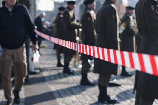 Soldiers in uniform stand behind a red-striped barricade during a military parade outdoors.