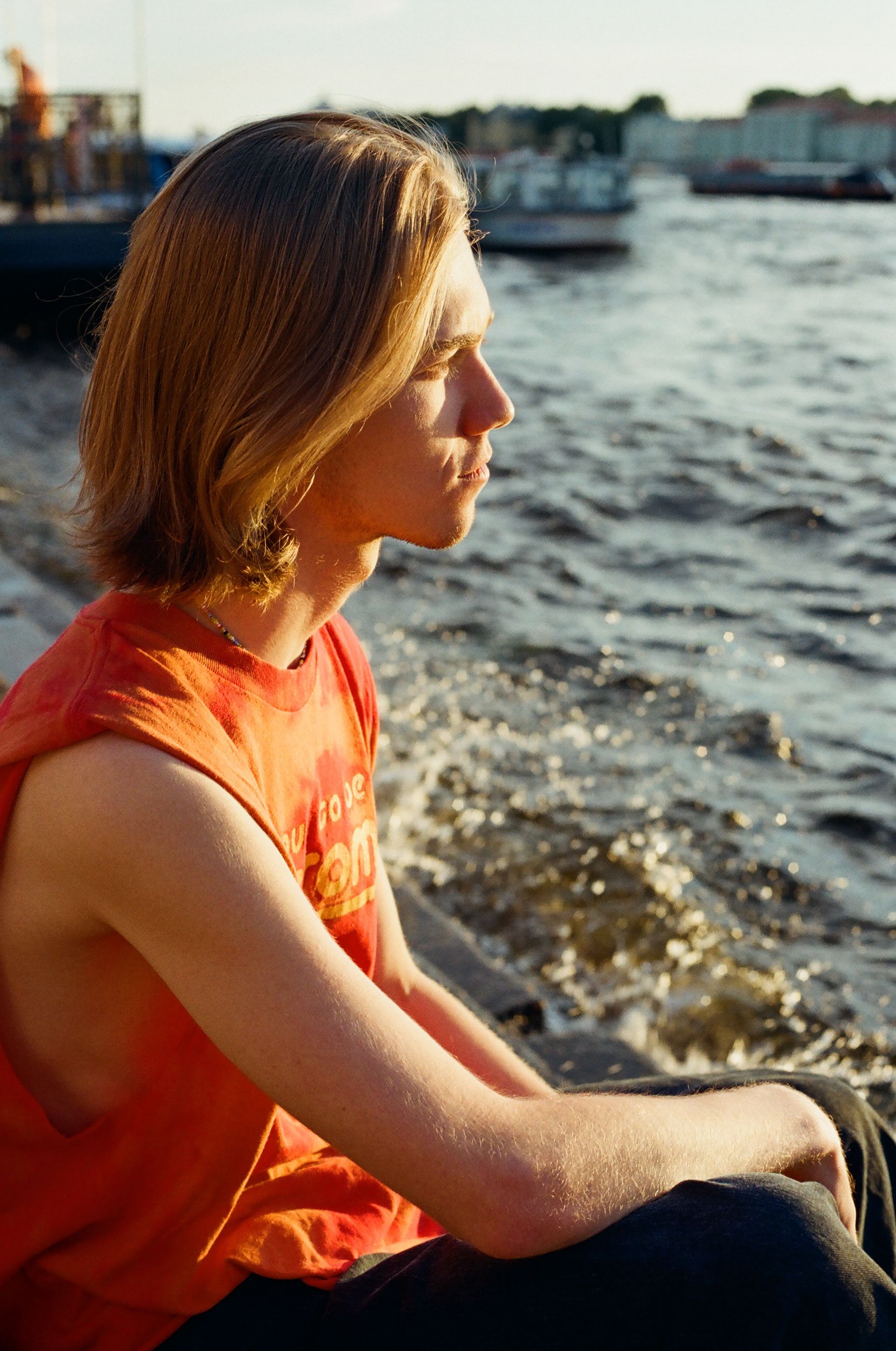 Side view of a young man with long hair sitting by the river during sunset, lost in thought.