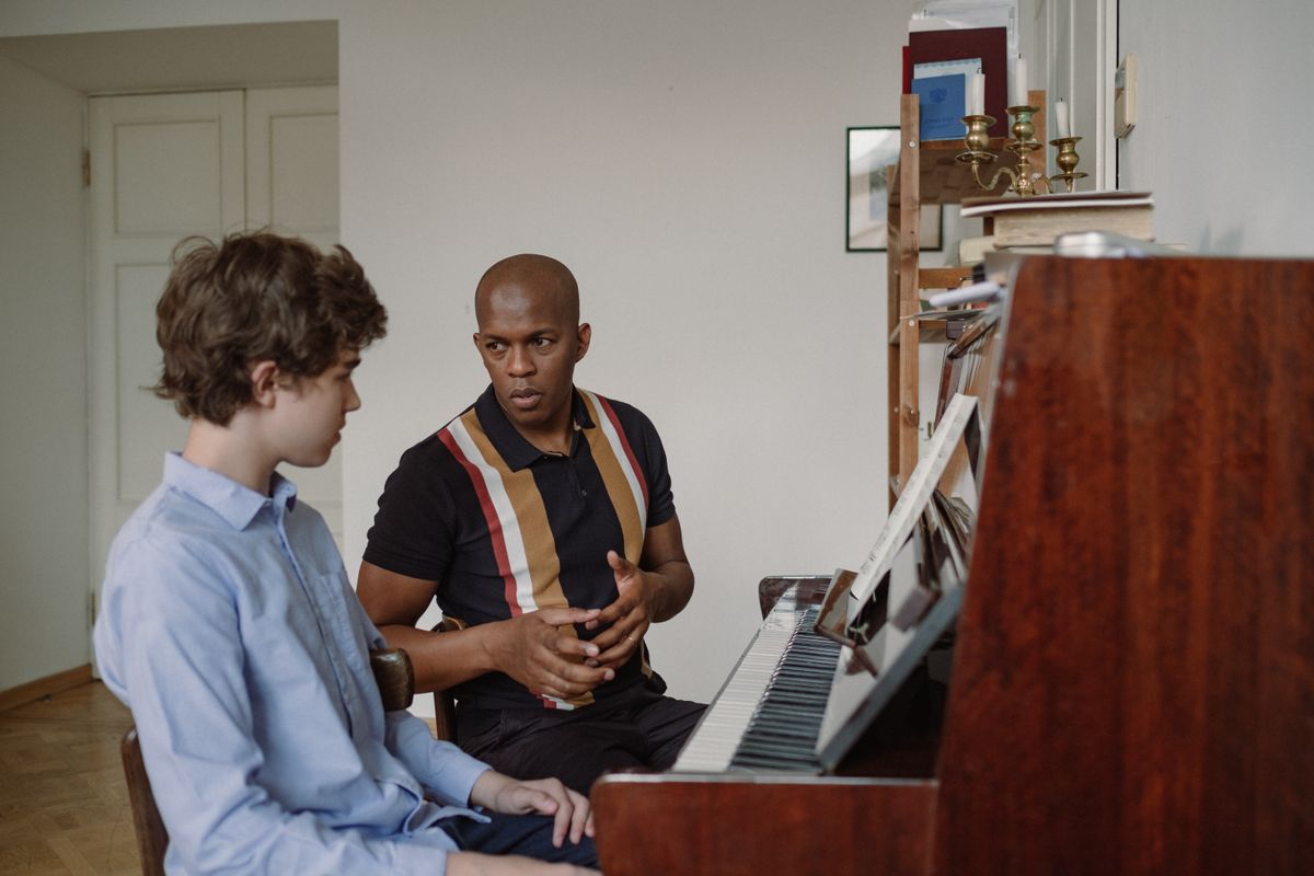 A teenage boy sitting at a piano while an adult beside him talks through the music