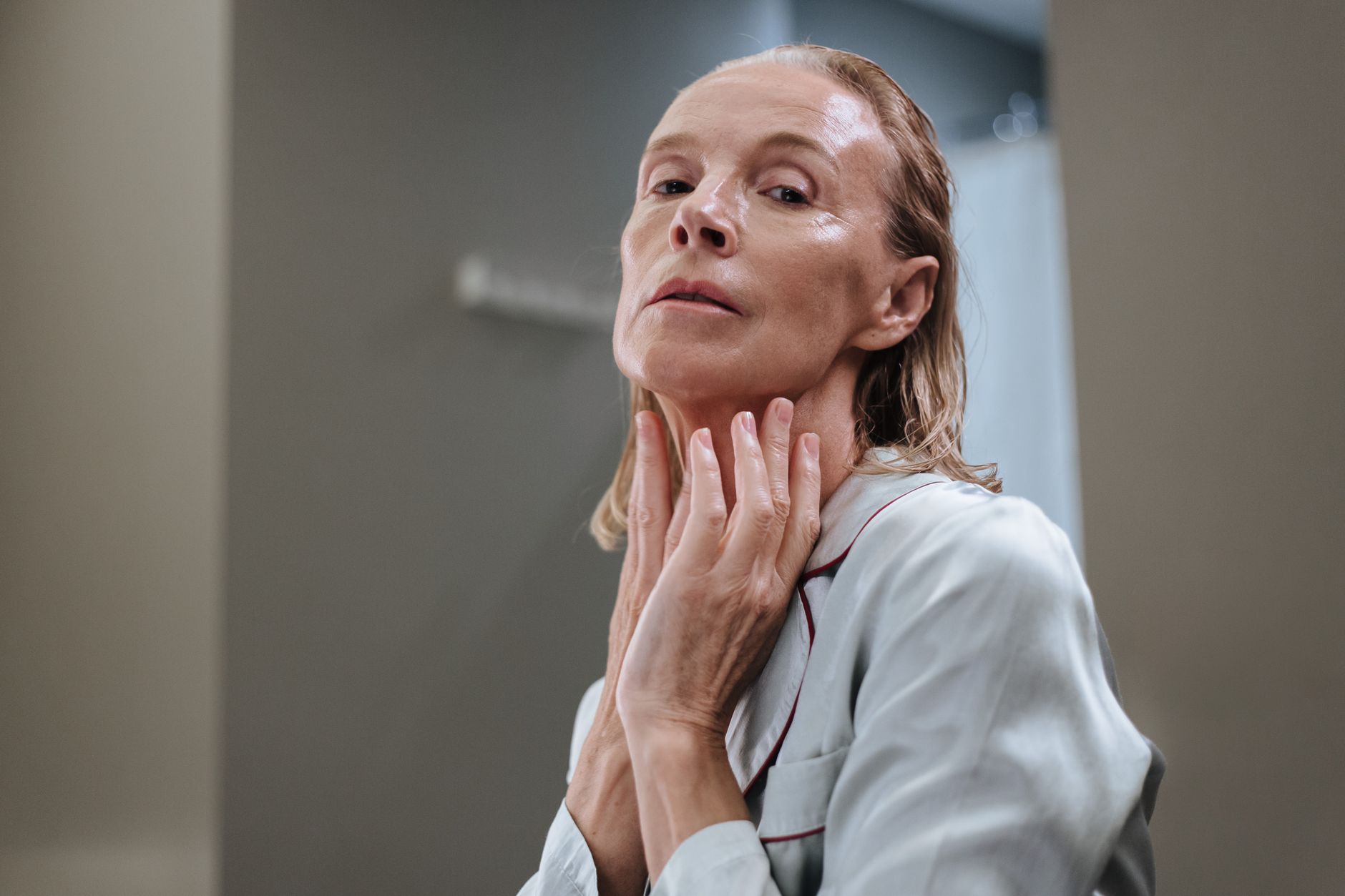 An elderly woman applying skincare product to her face in a bathroom setting.