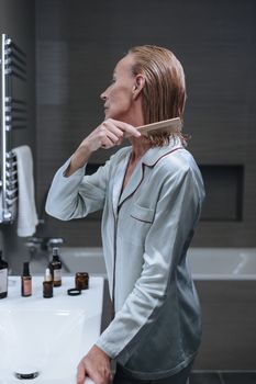 Senior woman combing hair in bathroom during morning self-care routine. Indoor setting with beauty products.