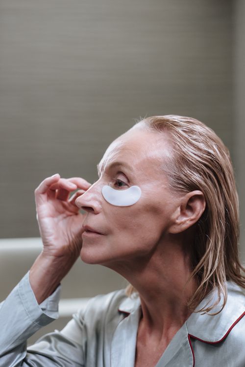 Free Elderly woman with blond hair applying under-eye skincare patch in a morning routine. Stock Photo