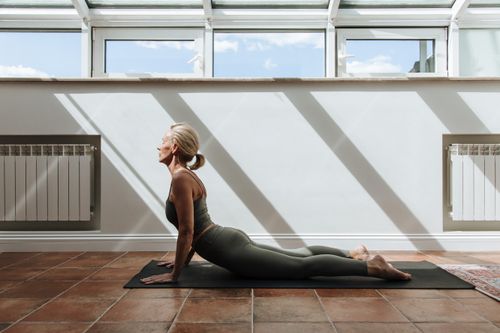 Free Senior woman performing yoga pose in a sunlit room, promoting healthy lifestyle and mindfulness. Stock Photo