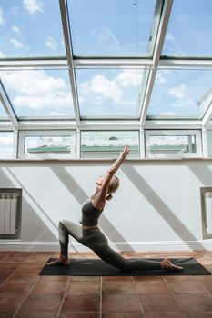 A woman in sportswear stretches on a yoga mat in a bright room with skylight.