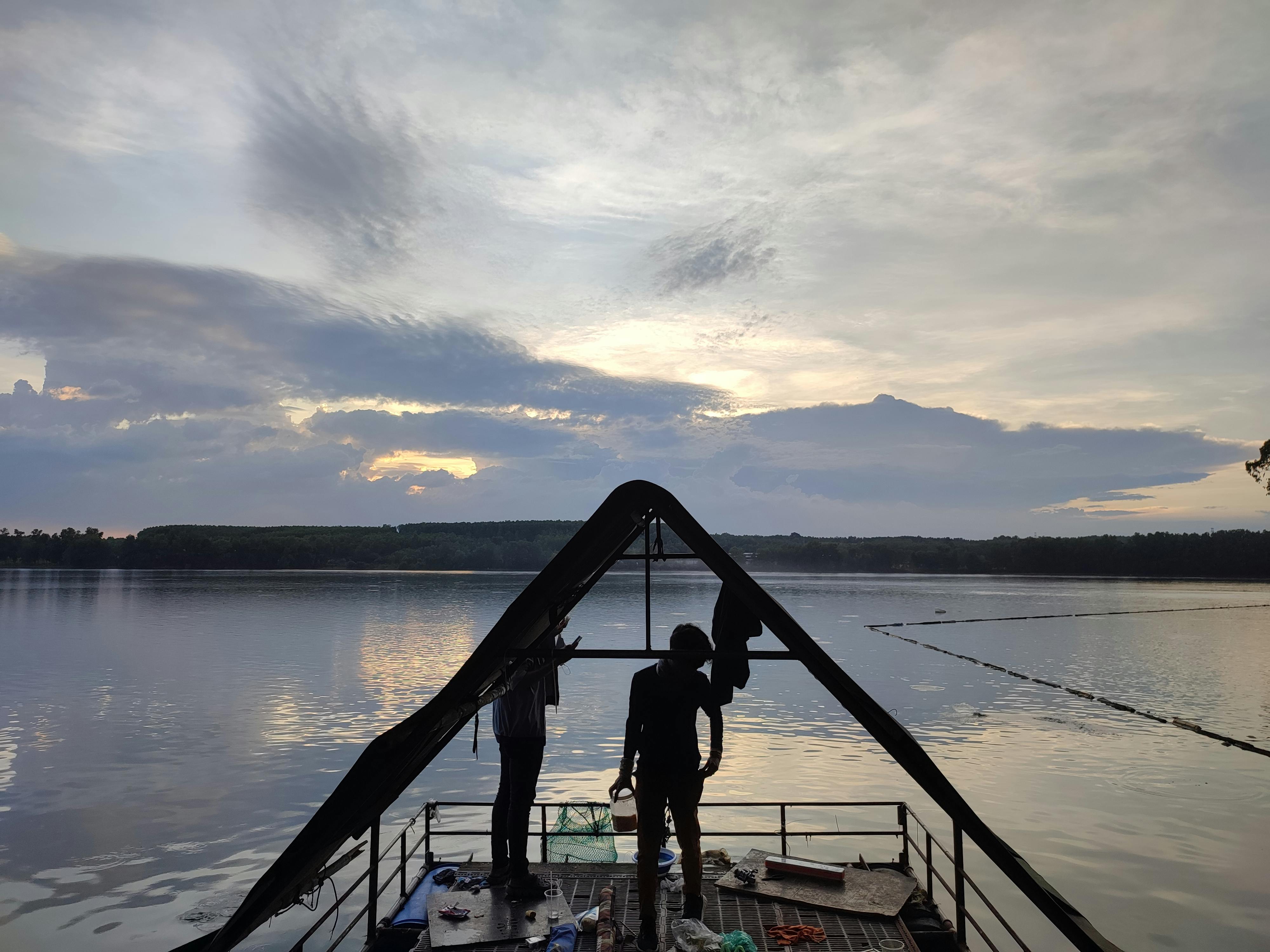 Silhouetted individuals stand on a wooden dock with a peaceful river and scenic sunset backdrop.